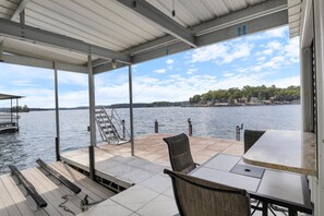 A covered outdoor deck with a table and chairs, overlooking a body of water and a distant shoreline. - Incredible Views, Spacious Deck, Private Dock, Gas Grill, Games & Cards (Gravois Mills)