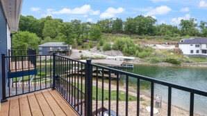 A wooden deck with a black railing overlooking a river, surrounded by greenery and a few buildings. - Our Cove Retreat - Perfect for Wave-free Swimming, Tanning & Prime Location Fun! (Lake Ozark)