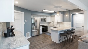 A modern kitchen with white cabinets, stainless steel appliances, a marble countertop, and a bar area with stools. - Our Cove Retreat - Perfect for Wave-free Swimming, Tanning & Prime Location Fun! (Lake Ozark)