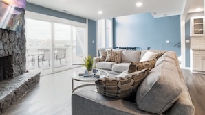 A modern living room with a stone fireplace, a large sofa, and a glass coffee table. - Our Cove Retreat - Perfect for Wave-free Swimming, Tanning & Prime Location Fun! (Lake Ozark)