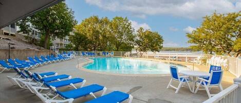 A pool area with blue lounge chairs and a table, surrounded by trees and a clear sky.