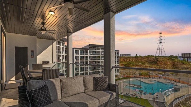 A covered outdoor area with a sofa and chairs, overlooking a swimming pool and a building with multiple balconies.