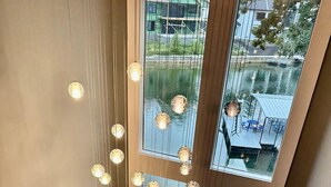 Ein modernes Interieur mit Essbereich, der einen Glastisch und Stühle, eine Hängeleuchte sowie große Fenster mit Blick auf ein grünes Gebäude und einen Steg umfasst.