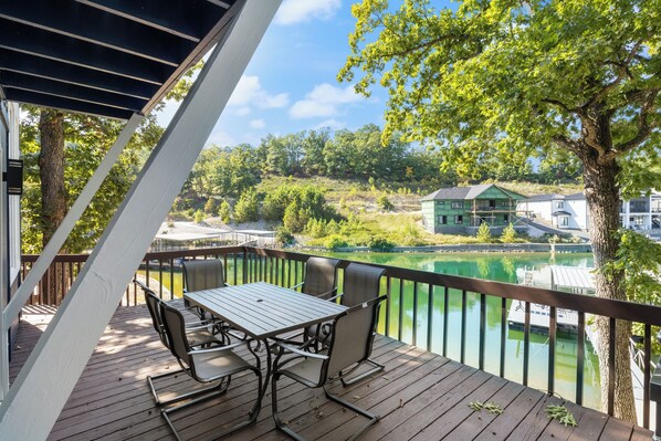 Een houten terras met een tafel en stoelen, met uitzicht op een waterpartij en een kas op de achtergrond.