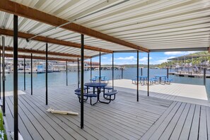 Een overdekt terras buiten met tafels en stoelen, met uitzicht op een jachthaven met boten en een verre kustlijn.