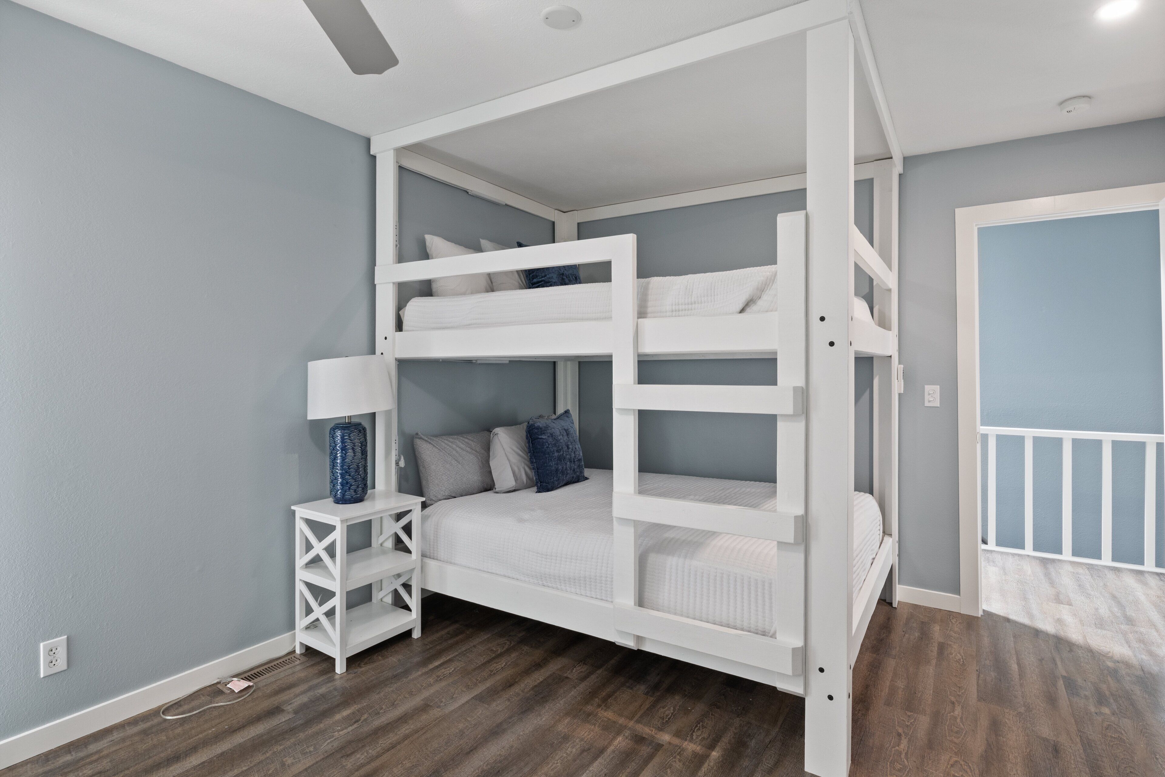 A bunk bed room with a white frame, a small white nightstand with a lamp, and a blue vase.
