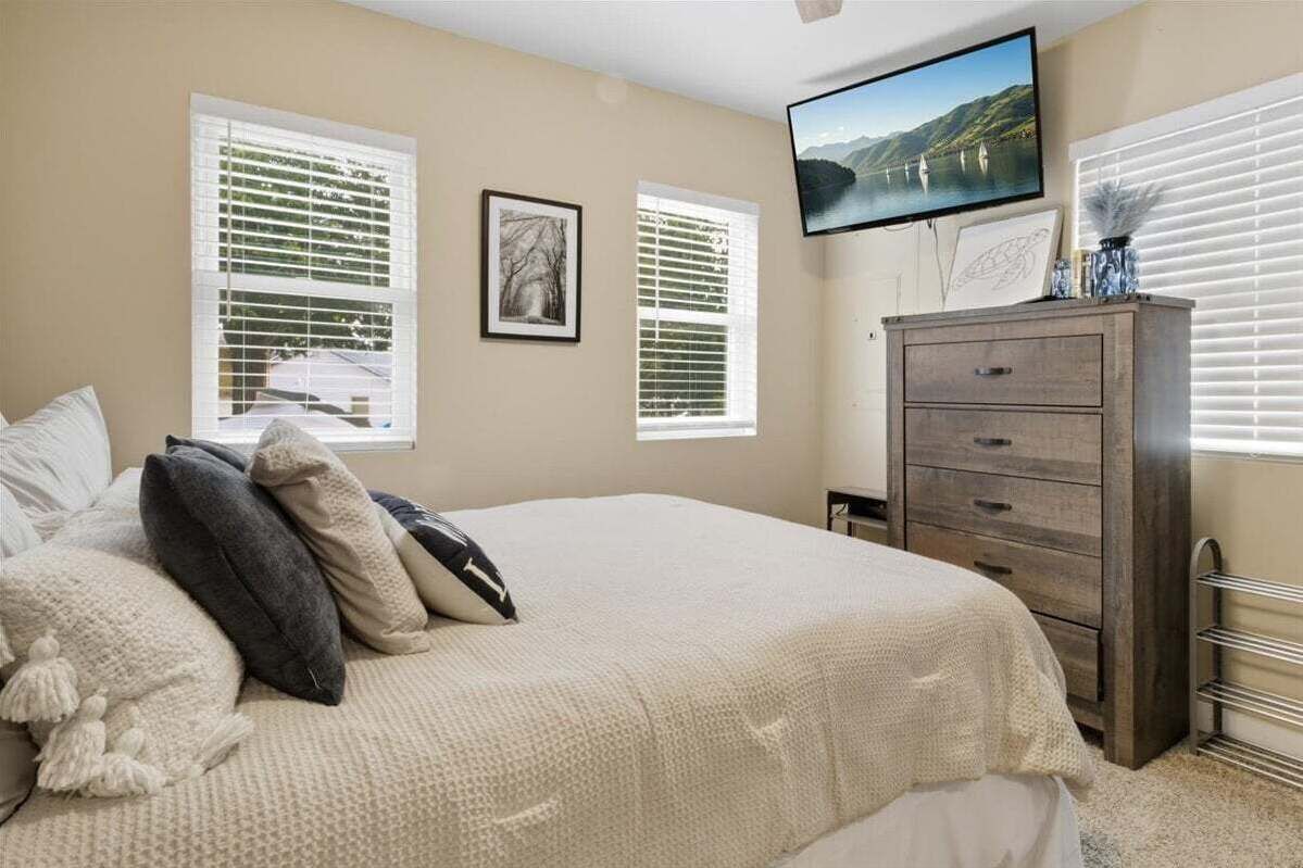 Une chambre à coucher avec un lit, deux fenêtres, une commode et un téléviseur fixé au mur.