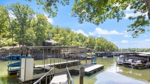 A docked boat with a covered area, a wooden pier, and a calm water body surrounded by trees. - Stunning Views of the 7.5mm, Private Dock, Spacious Decks, Kayaks & More! (Gravois Mills)