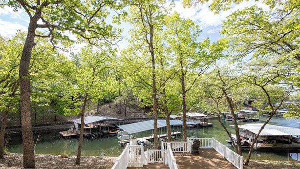 A wooden deck leading to a dock area with boats and covered structures by a calm water body, surrounded by trees. - Stunning Views of the 7.5mm, Private Dock, Spacious Decks, Kayaks & More! (Gravois Mills)