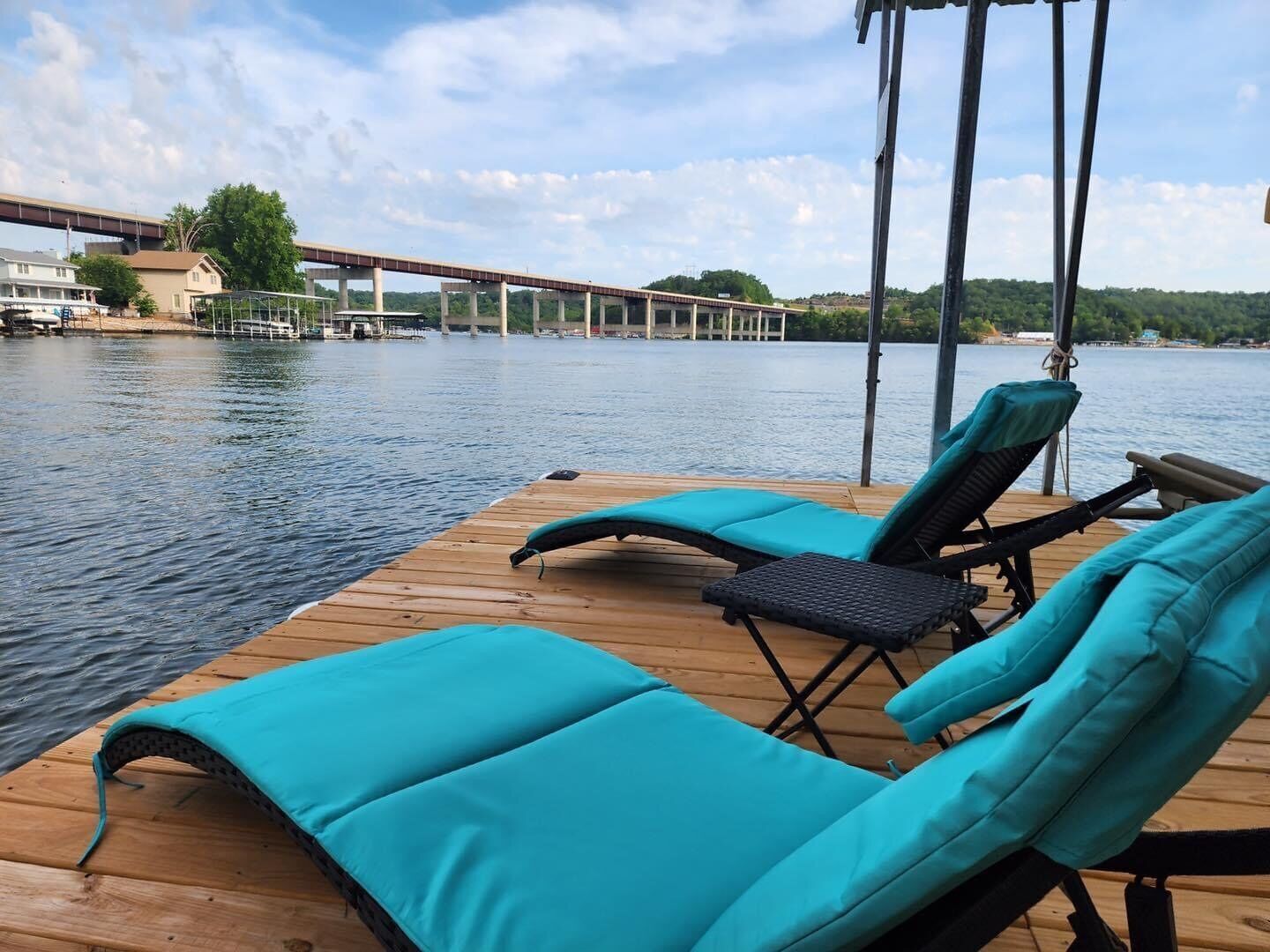 Une terrasse en bois avec deux chaises longues et une petite table, donnant sur une étendue d’eau calme, avec un pont en arrière-plan.