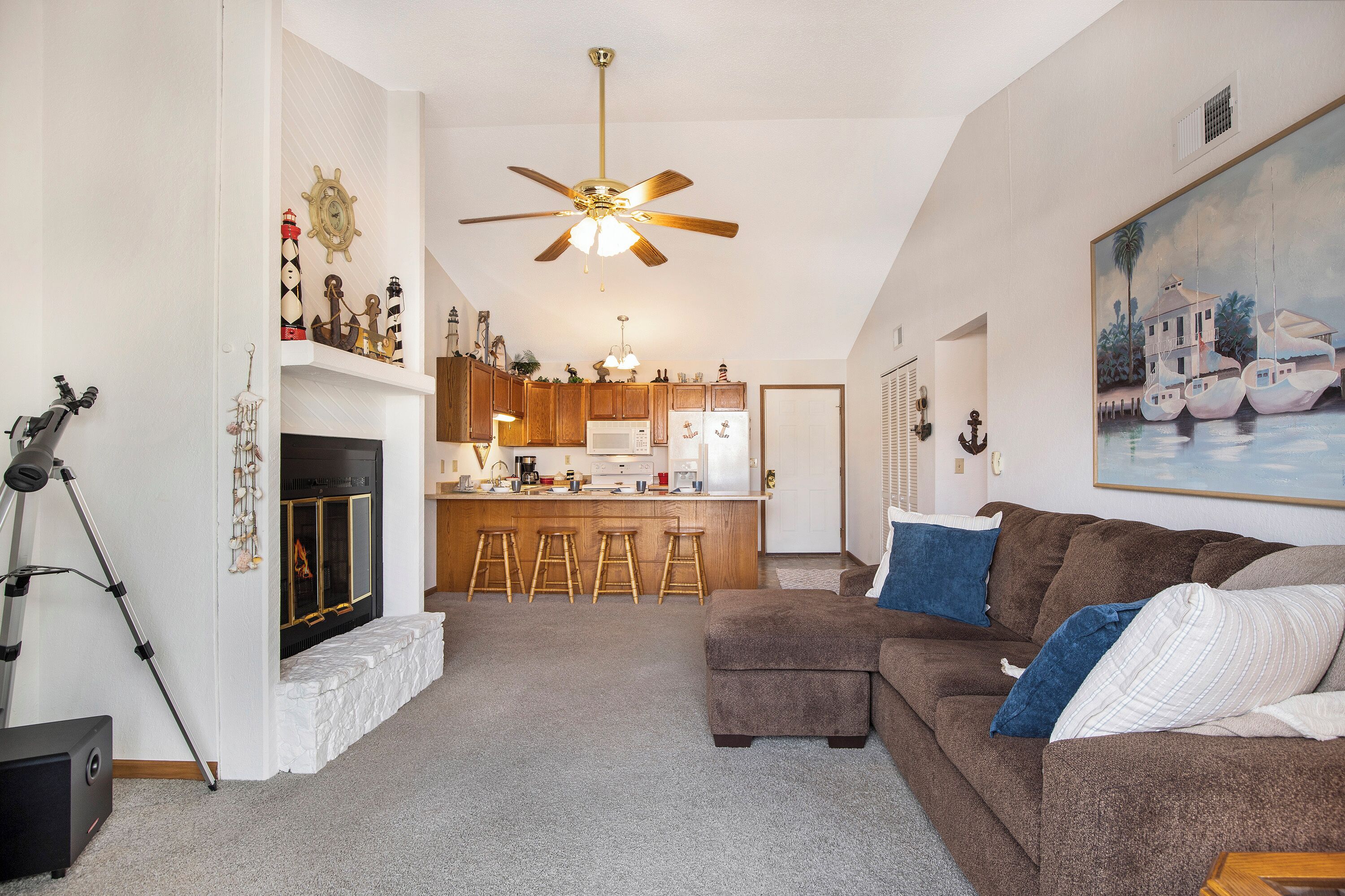 A living room with a fireplace, a ceiling fan, and a kitchen in the background.