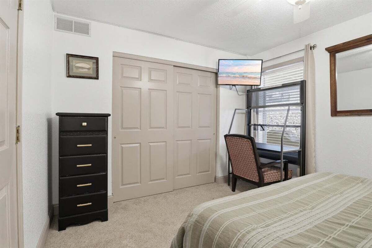 A bedroom with a bed, a desk with a chair, a dresser, a TV, and a window with blinds.