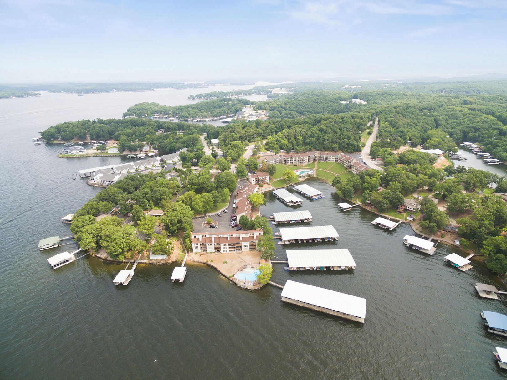 An aerial view of a lakeside property with multiple docked boats, a central building with a swimming pool, and surrounding greenery.