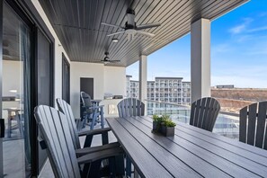 A modern outdoor dining area with a wooden table and chairs, a ceiling fan, and a view of buildings in the background.