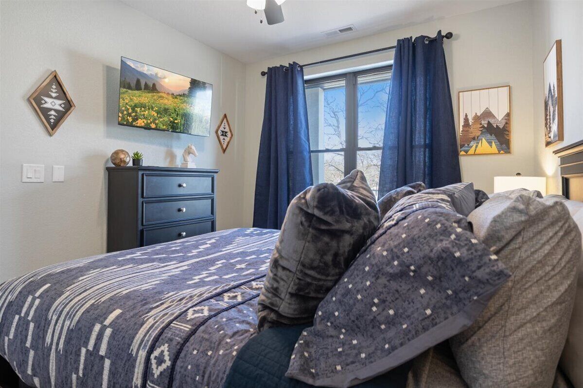 A bedroom with a bed, a dresser, a TV on the wall, and a window with curtains.