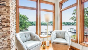 A room with two grey armchairs, a floor lamp, and large windows overlooking a river. - Amazing Cove, Fenced Yard, & Game Room! (Sunrise Beach)
