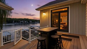 A wooden deck with a railing, a table, and two chairs, overlooking a body of water with boats and a dock in the background. - Amazing Cove, Fenced Yard, & Game Room! (Sunrise Beach)