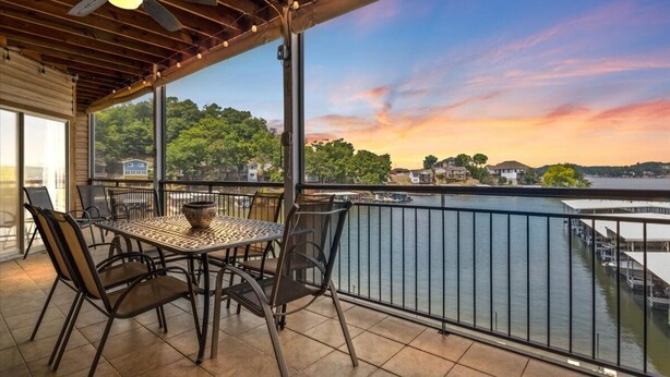 A covered outdoor patio with a table and chairs, overlooking a body of water and a residential area during sunset. - Lakefront 1st-floor Condo, Screened Patio, Pools & Courts (Lake Ozark)