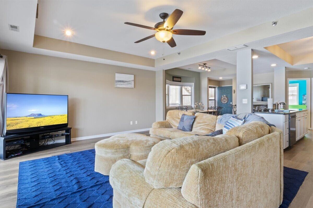 A living room with a large flat-screen TV, beige sofas, a ceiling fan, and a kitchen area in the background.