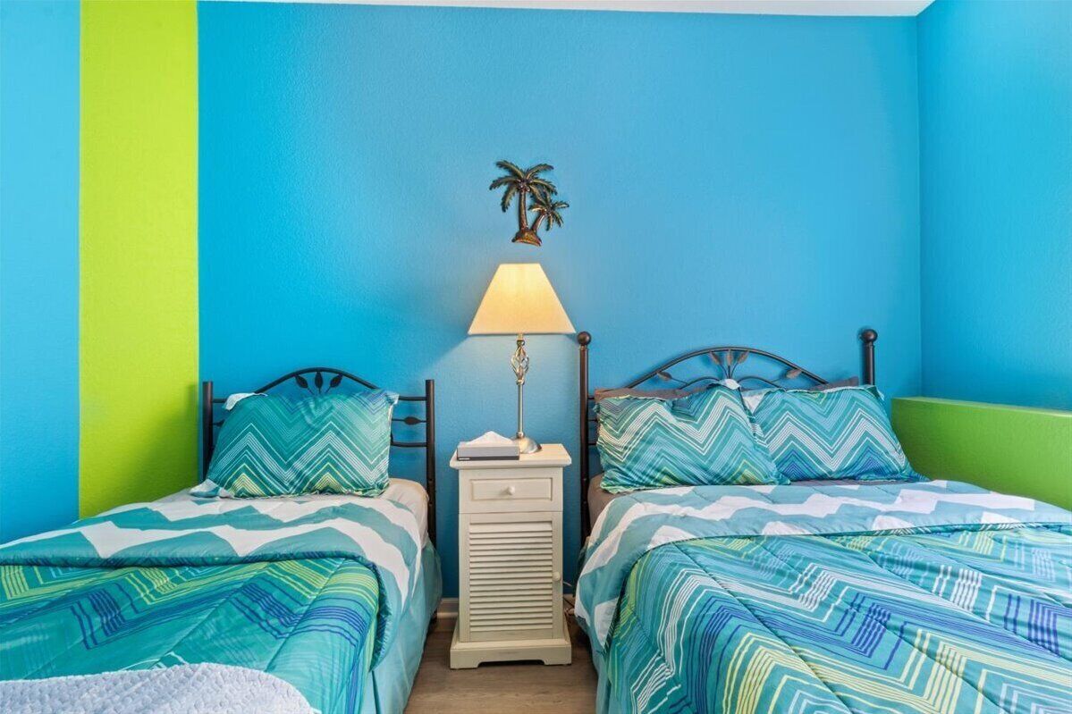 A room with two beds, each with patterned bedding, a nightstand with a lamp, and a wall-mounted palm tree decoration.
