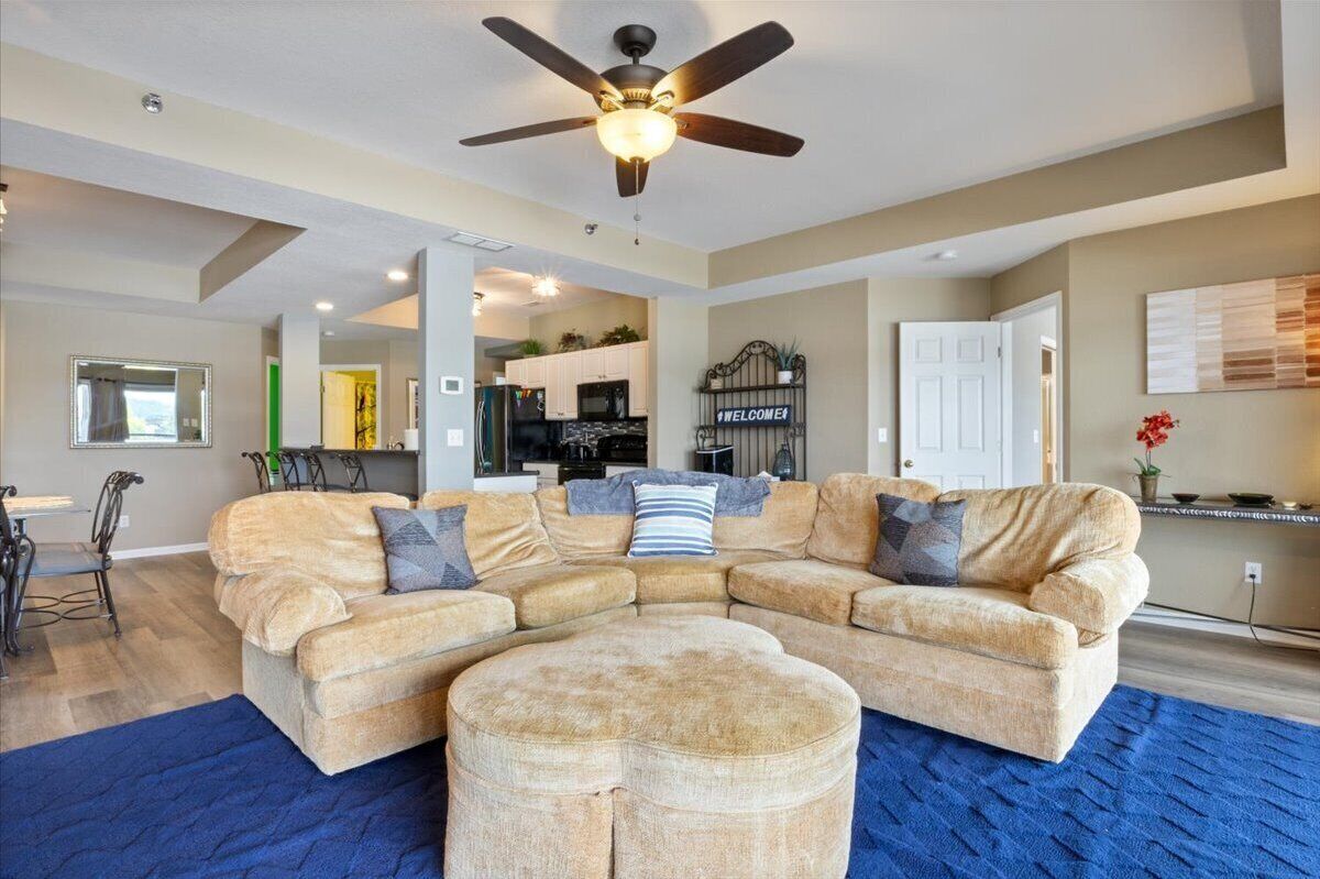 A modern living room with a sectional sofa, ottoman, ceiling fan, and a view into the kitchen.