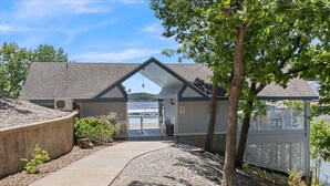 A building with a sloped roof, a gravel pathway leading to a gated entrance, and a view of a body of water in the background. - Games, Lake Views, Ease of Access to Unit & Attractions, Family-friendly, Outdoor Comfort (Lake Ozark)