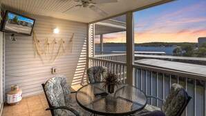 A balcony with a glass table, chairs, a ceiling fan, and a TV mounted on the wall. - Games, Lake Views, Ease of Access to Unit & Attractions, Family-friendly, Outdoor Comfort (Lake Ozark)