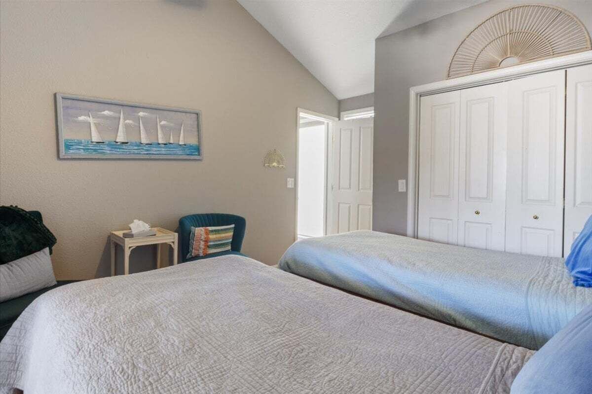 A bedroom with two beds, a chair, a small table, and a painting of sailboats on the wall.