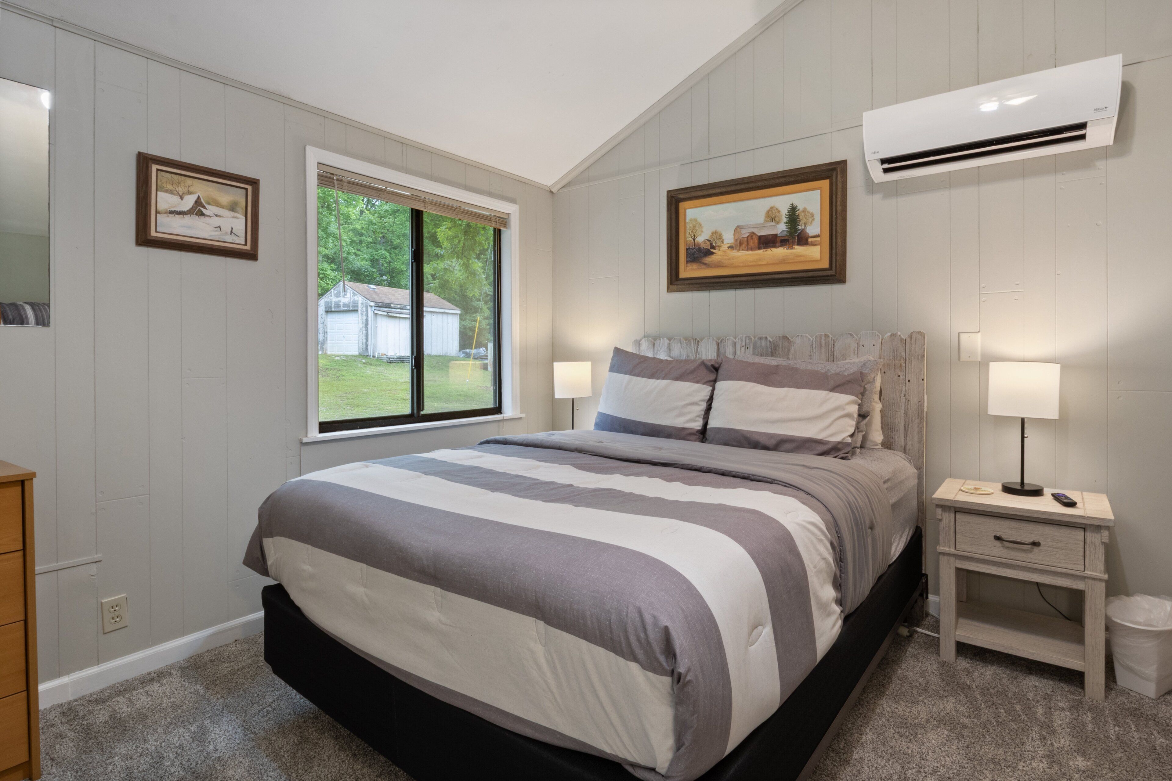 Une chambre avec un grand lit, une fenêtre donnant sur une maison, un climatiseur et une table de chevet avec une lampe.