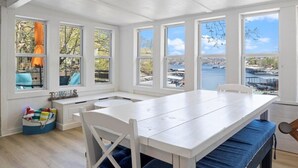 A dining area with a white table and chairs, large windows with a view of a marina, and a bench with a blue cushion.
