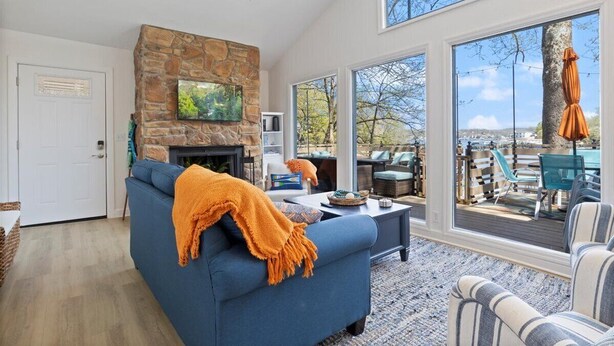 A modern living room with a stone fireplace, large windows, and outdoor seating visible through the glass doors.