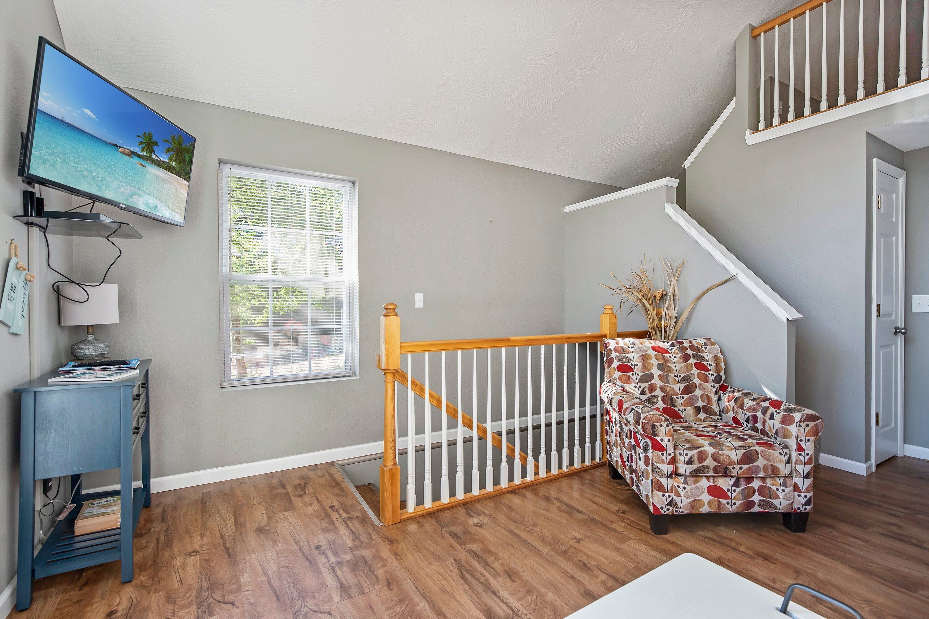 A living room with a wooden floor, a television mounted on the wall, a patterned armchair, and a staircase with a white railing.