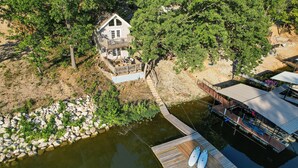A house with a dock extending into a body of water, surrounded by trees and a rocky shoreline. - Private Dock, Paddle Boards, Views & Hot Spots! (Osage Beach)
