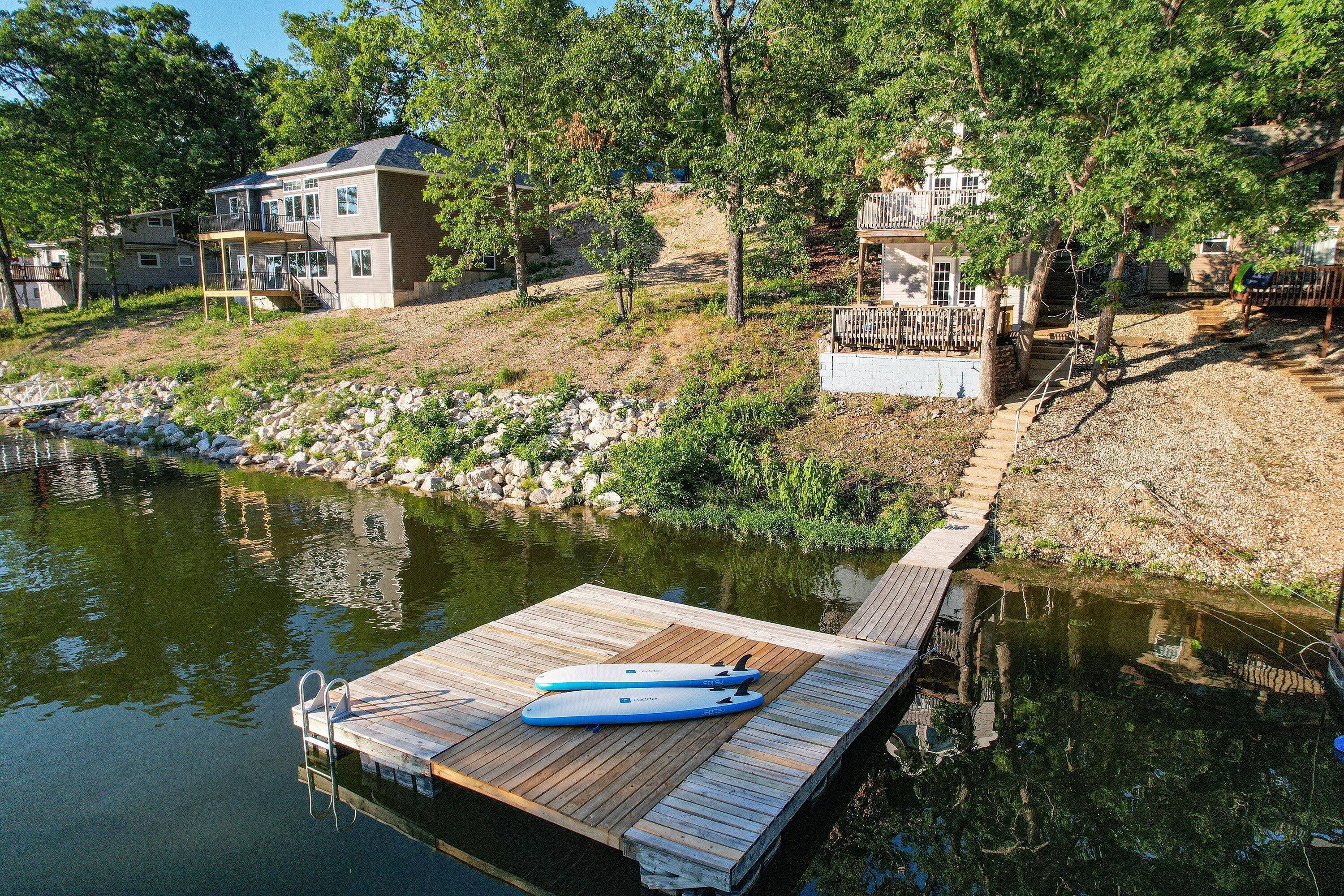 Un muelle de madera con un kayak y una escalera, rodeado por un lago tranquilo, con casas y árboles al fondo.