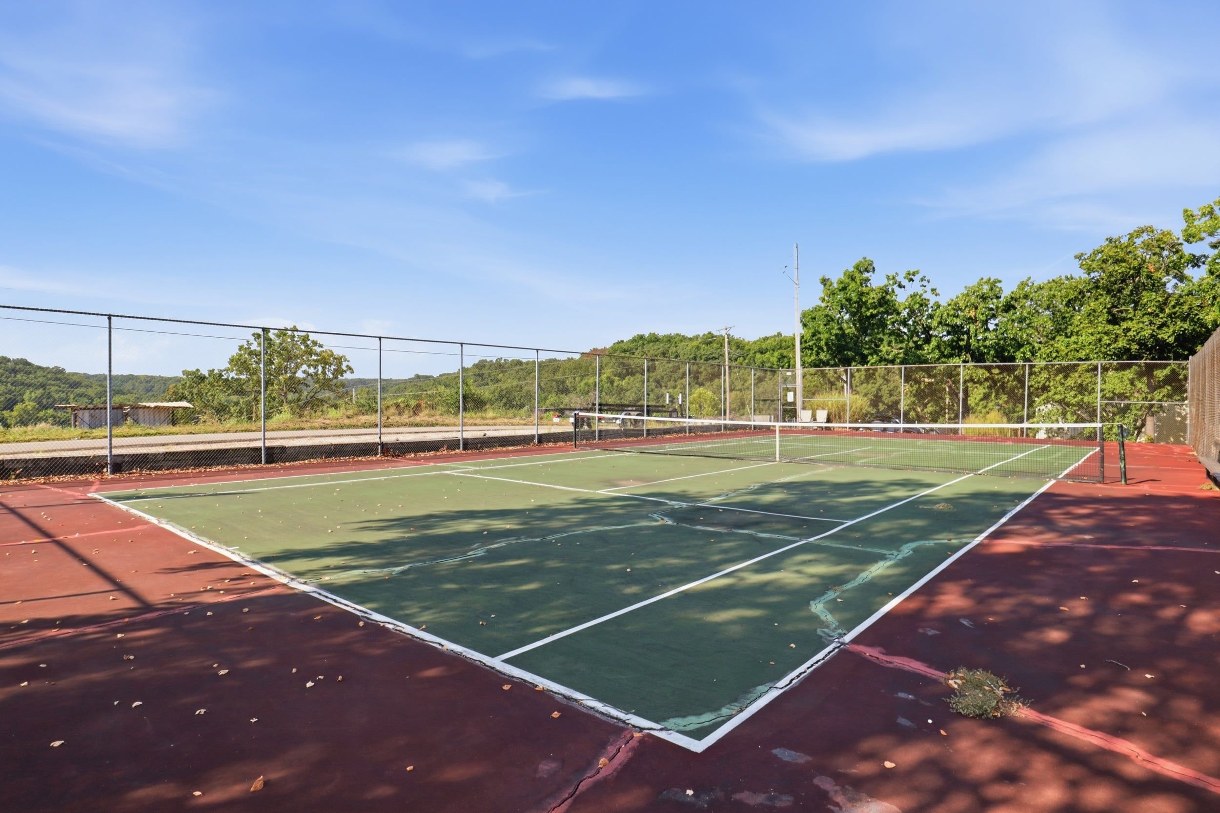 An outdoor tennis court with a green playing surface and red surrounding area, surrounded by a chain-link fence and trees.