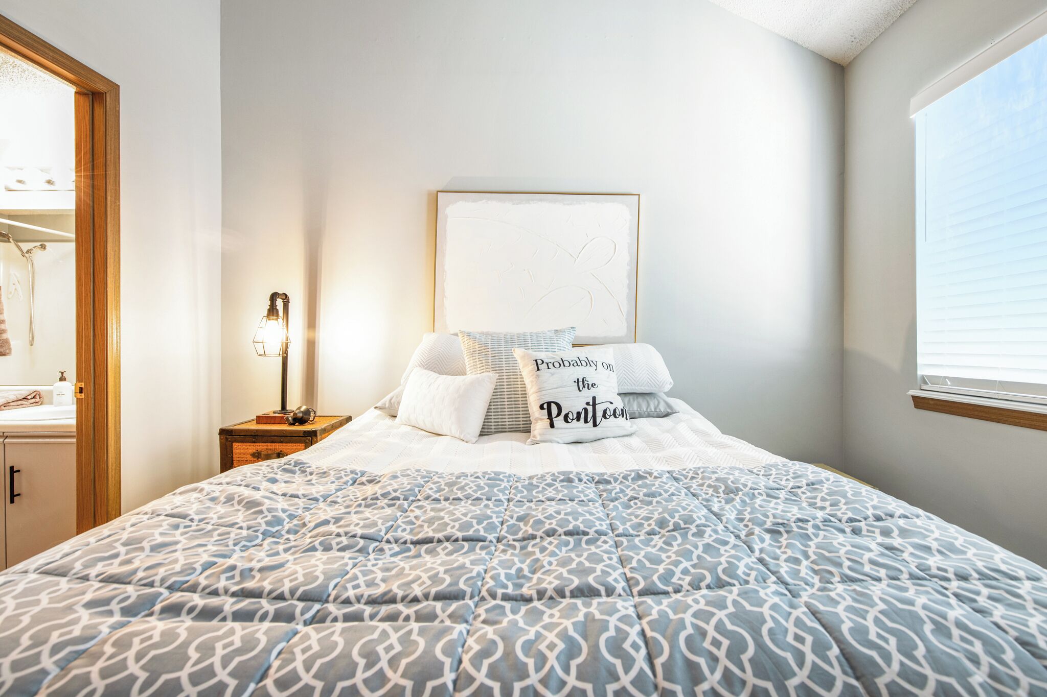 Un dormitorio con cama, una mesita de noche con lámpara, una obra de arte enmarcada en la pared y una ventana con persianas.
