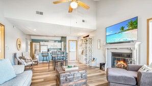 A spacious living room with a fireplace, a television displaying a beach scene, and a kitchen in the background. - Poolside Condo With Amazing Lake Views & Design (Osage Beach)