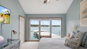 A bedroom with a large bed, a glass table, a chair, a TV, and a view of a lake and dock. - Poolside Condo With Amazing Lake Views & Design (Osage Beach)