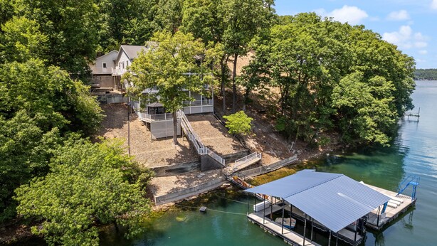 A lakeside property with a dock, a covered boat launch, and a house surrounded by dense trees. - Amazing Game Room With Dock Tram to Huge Dock - Great For Families (Lake Ozark)