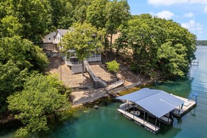 A lakeside property with a dock, a covered boat launch, and a house surrounded by dense trees. - Amazing Game Room With Dock Tram to Huge Dock - Great For Families (Lake Ozark)