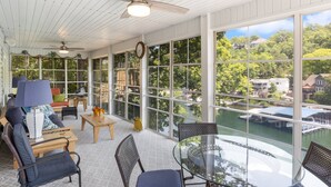 A sunlit room with a view of a lake, furnished with a sofa, chairs, and a glass table. - Amazing Game Room With Dock Tram to Huge Dock - Great For Families (Lake Ozark)