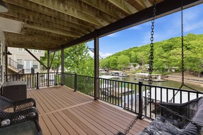 Une terrasse en bois, meublée avec des meubles en osier, un balancoir et une vue sur un lac où naviguent des bateaux.