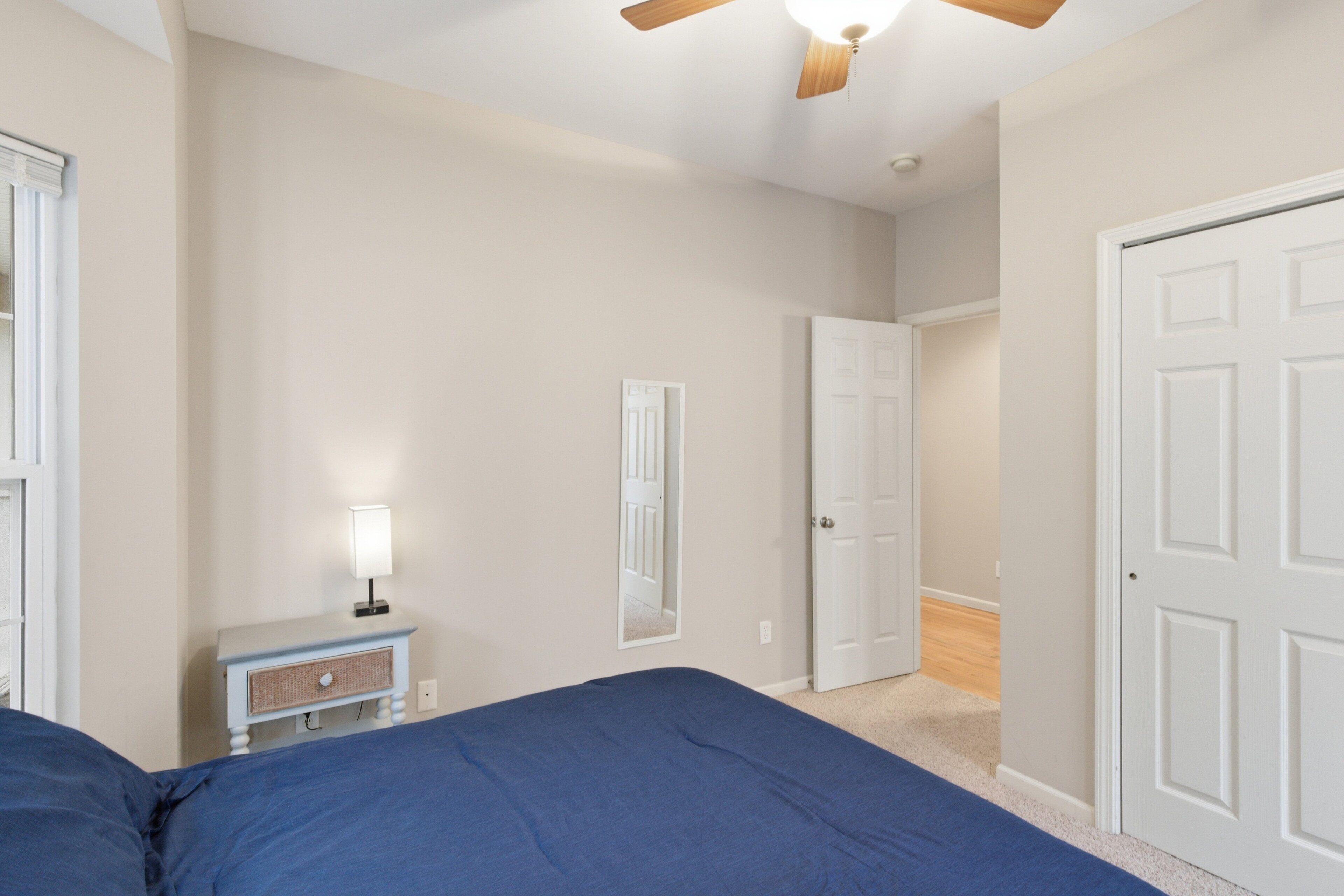 Quarto com cama, mesa de cabeceira com abajur, ventilador de teto e porta para outro cômodo.