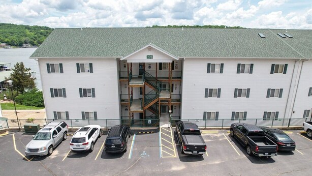 A multi-story building with a central staircase, parking lot with various vehicles, and a view of a lake. - Boat Slip, 3 Pools, Golf, Ozarks Ampitheater, & Amazing Lake Views at Big Niangua (Camdenton)