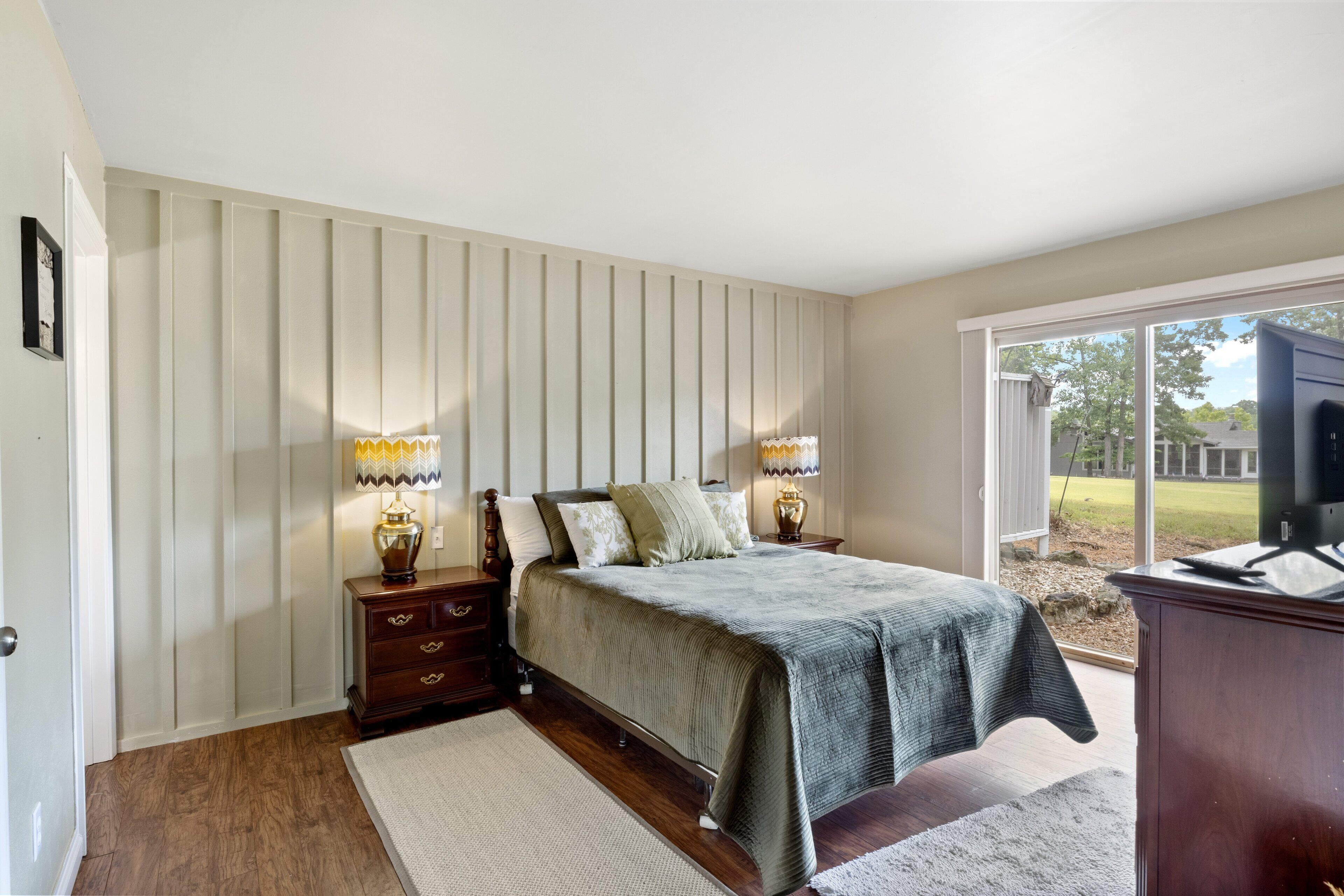 A bedroom with a large bed, two bedside tables with lamps, a wooden dresser, a large window with a view of the outdoors, and a door on the left.
