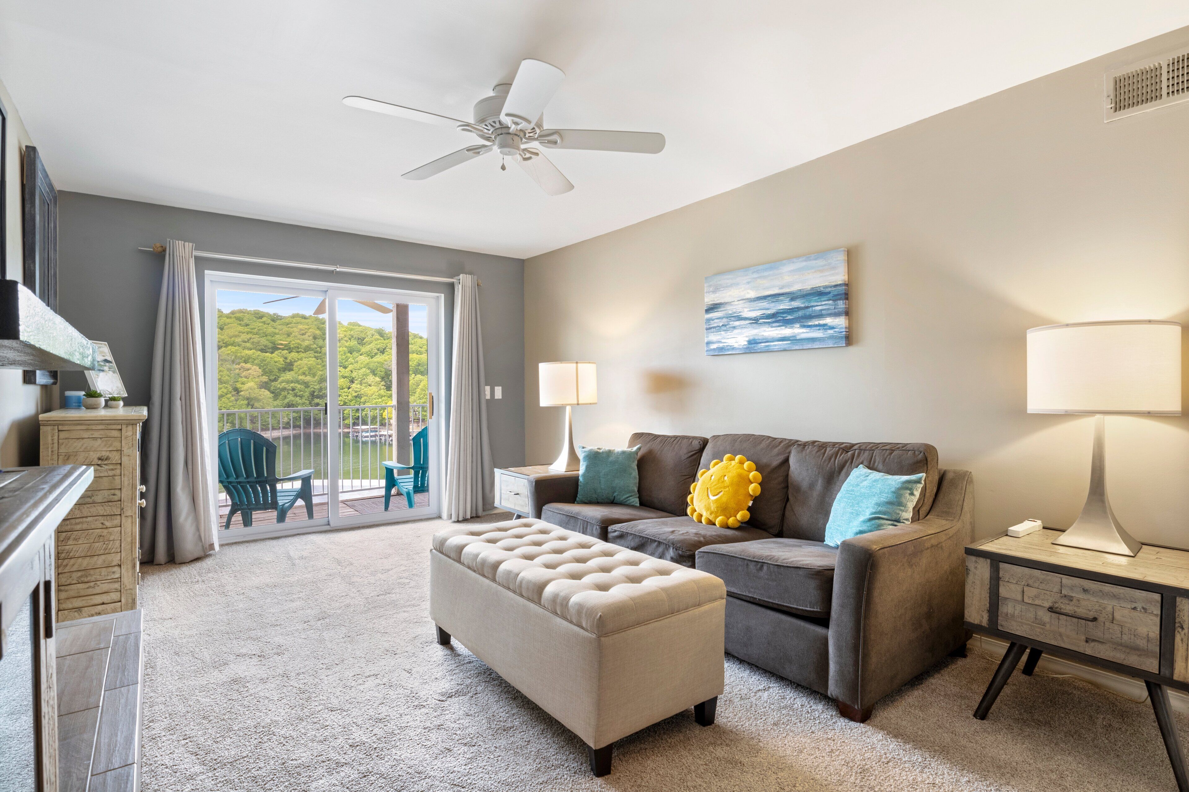 A living room with a sofa, ottoman, and a ceiling fan. There is a sliding glass door leading to a balcony with chairs.