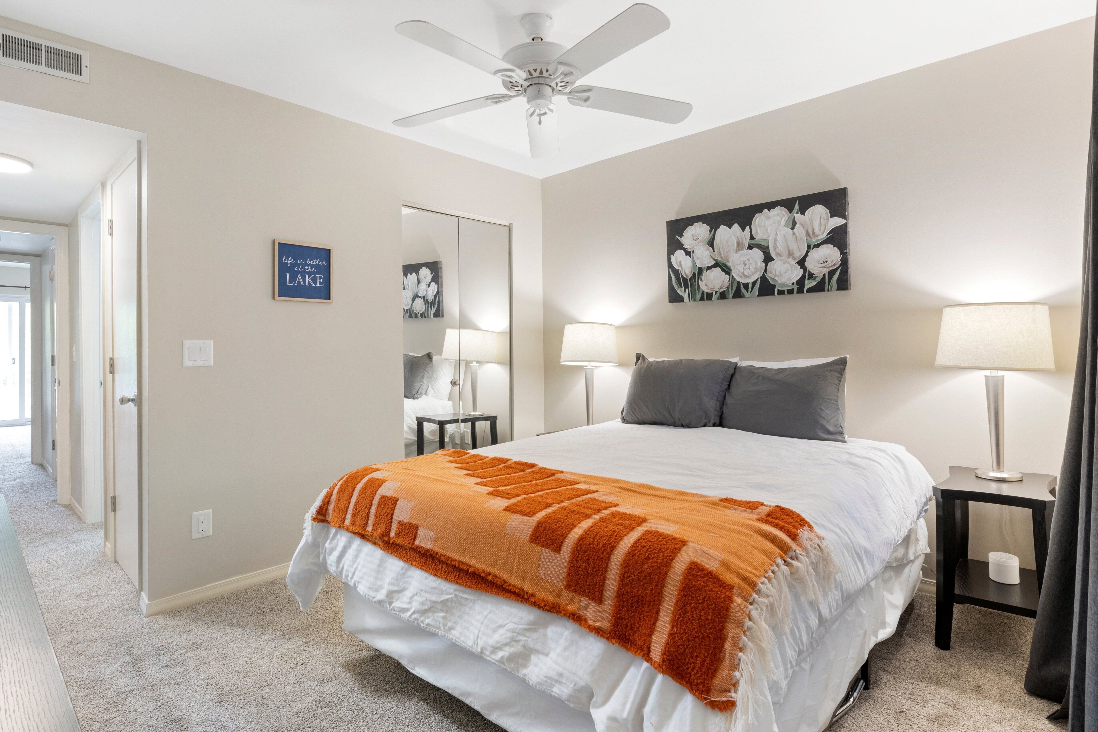 Une chambre à coucher avec un lit, deux lampes de chevet, un ventilateur de plafond, un miroir et un tableau représentant des fleurs.