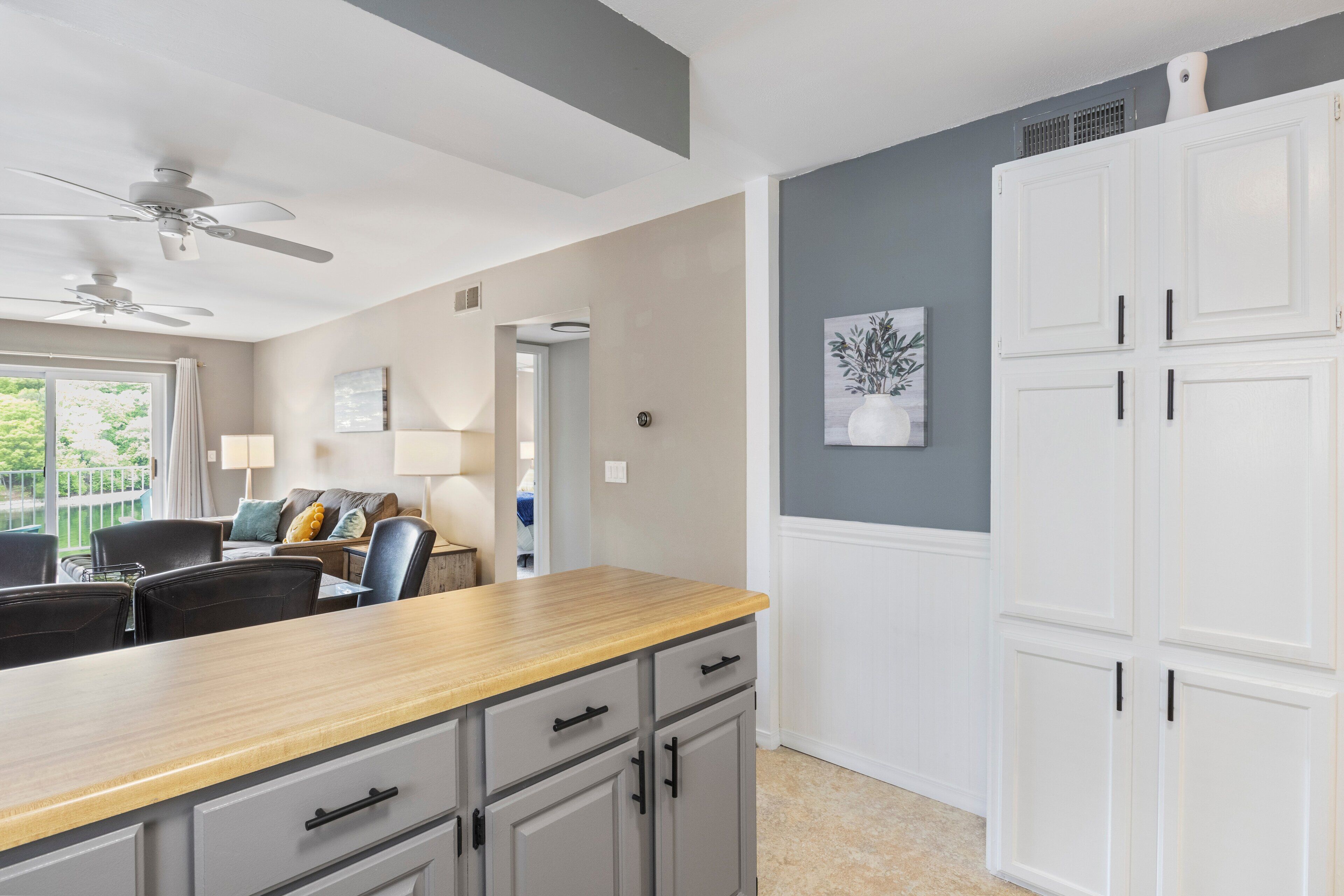 A modern kitchen with a wooden countertop, grey cabinets, and white upper cabinets. There is a ceiling fan, a dining area with chairs, and a view of the outdoors.