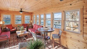 A cozy cabin interior with wooden walls, large windows, a ceiling fan, and a round table with chairs. - Game Room, Dock Life, Fire Pit & Hot Tub! (Lake Ozark)