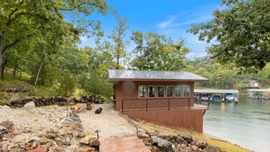 A wooden cabin with a metal roof, a dock, and a rocky shoreline. - Game Room, Dock Life, Fire Pit & Hot Tub! (Lake Ozark)